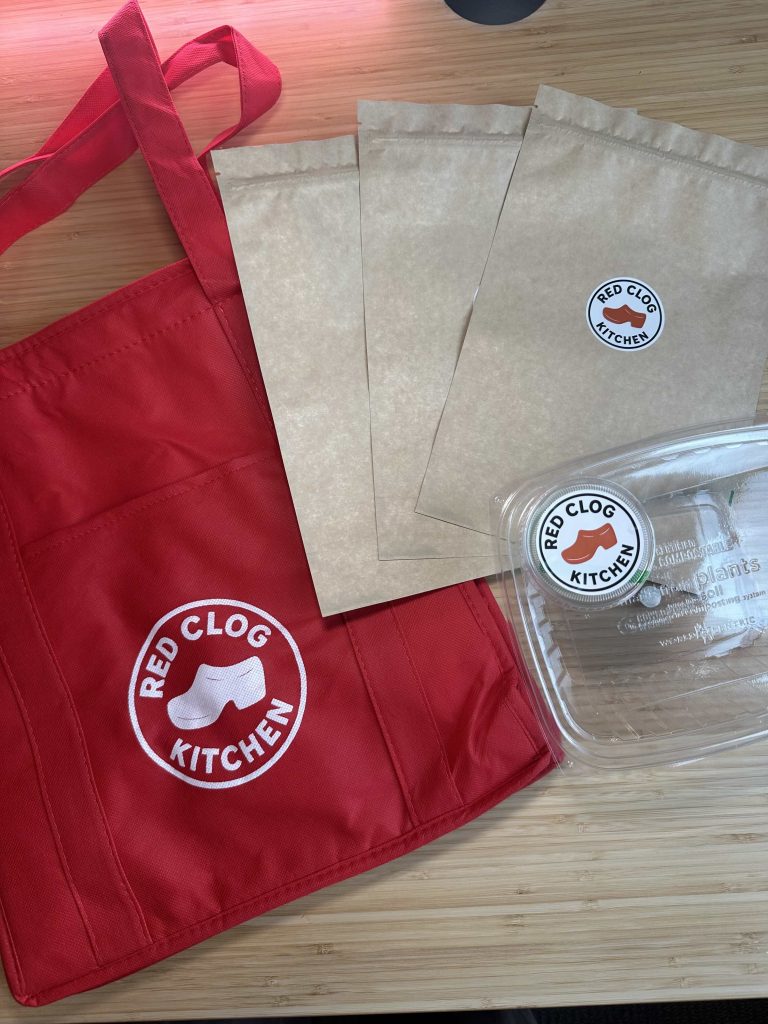 Red Clog Kitchen's sustainable packaging, including compostable containers, and a reusable fabric bag.